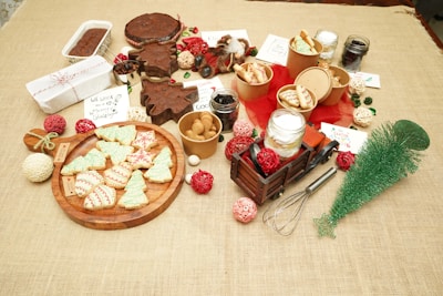 A variety of cookies and Christmas-themed treats are arranged on a table, surrounded by festive decorations like a small green tree, red and white ornaments, and packaged gift items. There are green and red frosted cookies shaped like trees and baubles, a whisk, jars filled with different ingredients, and a toy wooden truck. Labels and handwritten notes are scattered throughout, adding to the holiday theme.