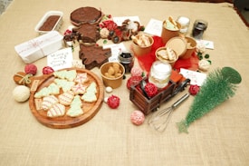 A variety of cookies and Christmas-themed treats are arranged on a table, surrounded by festive decorations like a small green tree, red and white ornaments, and packaged gift items. There are green and red frosted cookies shaped like trees and baubles, a whisk, jars filled with different ingredients, and a toy wooden truck. Labels and handwritten notes are scattered throughout, adding to the holiday theme.