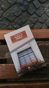 A book titled 'Hüzün ve Tesadüf' by Mustafa Kutlu is placed on a wooden bench with cobblestone pavement in the background. The book cover features an illustration of a window with vibrant red flowers.