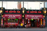 An exterior view of 'The Scottish Pub' featuring red awnings with the pub's name and neon signs displaying the name as well. There are outdoor seating arrangements with some patrons sitting at tables. The pub has a historic and inviting appearance, with classic lanterns and decorative plant arrangements above the entrance. The facade includes large windows reflecting the surroundings, possibly showing a distant church tower.