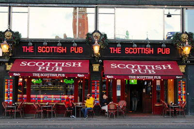 An exterior view of 'The Scottish Pub' featuring red awnings with the pub's name and neon signs displaying the name as well. There are outdoor seating arrangements with some patrons sitting at tables. The pub has a historic and inviting appearance, with classic lanterns and decorative plant arrangements above the entrance. The facade includes large windows reflecting the surroundings, possibly showing a distant church tower.