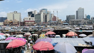 A busy urban market scene with numerous umbrellas in various colors providing shade over stalls and people. Tall buildings form the backdrop, indicating a metropolitan setting. The market area appears crowded, with many makeshift structures and vendors displaying their goods.