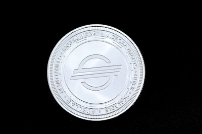 A silver coin featuring an emblem in the center with a pattern of text along the edge, set against a solid black background.
