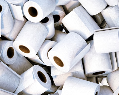 A large pile of white toilet paper rolls, each with a textured pattern. The rolls are scattered in a seemingly random manner, some stacked and others resting in different orientations with visible cardboard tubes.