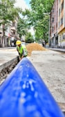 A construction worker wearing a yellow hard hat and high-visibility vest is engaged in roadwork, laying what appears to be a large blue pipe in a trench along an urban street. The scene takes place on a sunlit street with trees and multistory residential buildings lining both sides. A pile of gravel or dirt is visible in the background, indicating ongoing construction efforts.