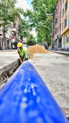 A construction worker wearing a yellow hard hat and high-visibility vest is engaged in roadwork, laying what appears to be a large blue pipe in a trench along an urban street. The scene takes place on a sunlit street with trees and multistory residential buildings lining both sides. A pile of gravel or dirt is visible in the background, indicating ongoing construction efforts.