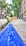 A construction worker wearing a yellow hard hat and high-visibility vest is engaged in roadwork, laying what appears to be a large blue pipe in a trench along an urban street. The scene takes place on a sunlit street with trees and multistory residential buildings lining both sides. A pile of gravel or dirt is visible in the background, indicating ongoing construction efforts.