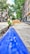 A construction worker wearing a yellow hard hat and high-visibility vest is engaged in roadwork, laying what appears to be a large blue pipe in a trench along an urban street. The scene takes place on a sunlit street with trees and multistory residential buildings lining both sides. A pile of gravel or dirt is visible in the background, indicating ongoing construction efforts.