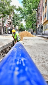 A construction worker wearing a yellow hard hat and high-visibility vest is engaged in roadwork, laying what appears to be a large blue pipe in a trench along an urban street. The scene takes place on a sunlit street with trees and multistory residential buildings lining both sides. A pile of gravel or dirt is visible in the background, indicating ongoing construction efforts.