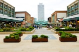 A bustling shopping precinct with various storefronts on either side, including a BHS store. People are walking and sitting in the open square area. Planters with greenery are arranged neatly along the wide pathway leading to a tall office building in the distance. The scene looks lively and modern.