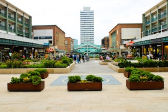 A bustling shopping precinct with various storefronts on either side, including a BHS store. People are walking and sitting in the open square area. Planters with greenery are arranged neatly along the wide pathway leading to a tall office building in the distance. The scene looks lively and modern.