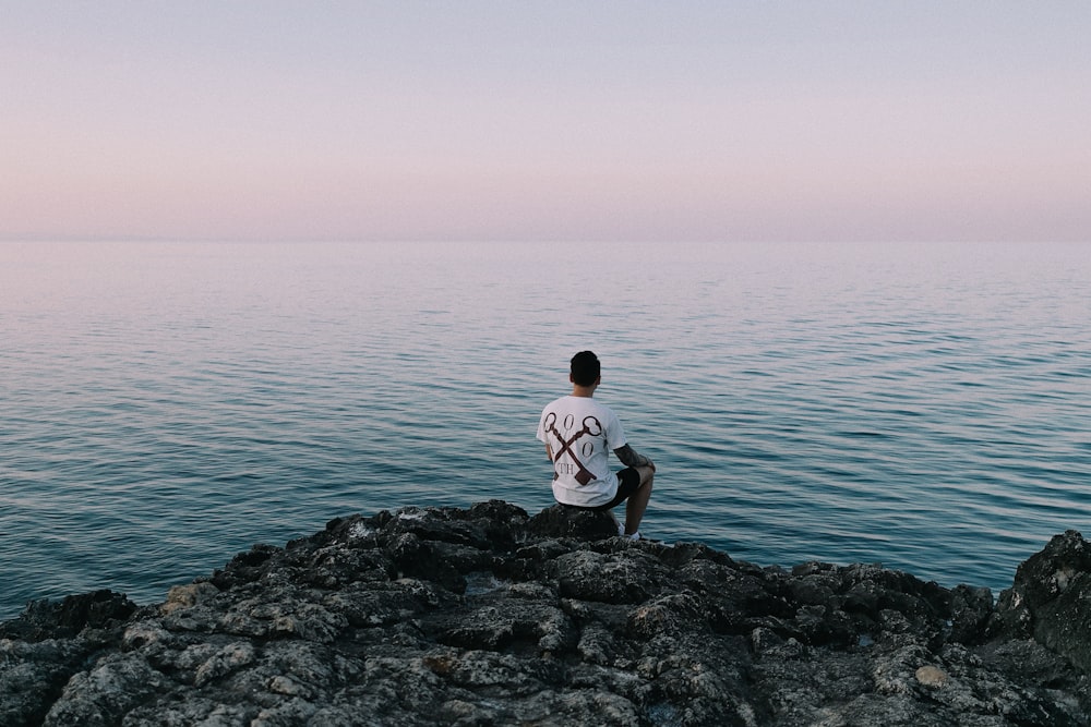 Hombre en camiseta blanca sentado en la roca junto al mar durante el día