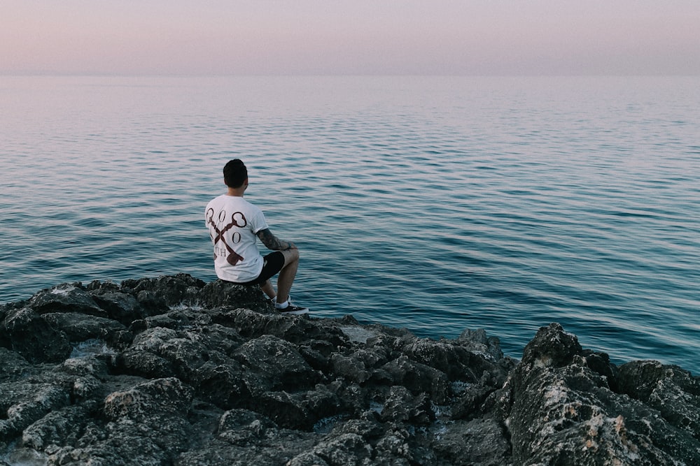 Hombre con camiseta blanca sentado en una roca gris cerca del cuerpo de agua durante el día