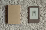 A closed beige hardcover book lies next to an e-reader with a low battery icon displaying a frowning face. Both items are placed on a textured, gray carpet.
