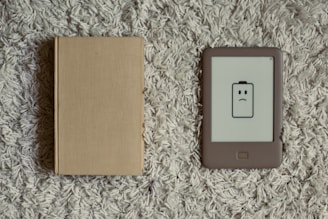 A closed beige hardcover book lies next to an e-reader with a low battery icon displaying a frowning face. Both items are placed on a textured, gray carpet.