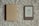 A closed beige hardcover book lies next to an e-reader with a low battery icon displaying a frowning face. Both items are placed on a textured, gray carpet.