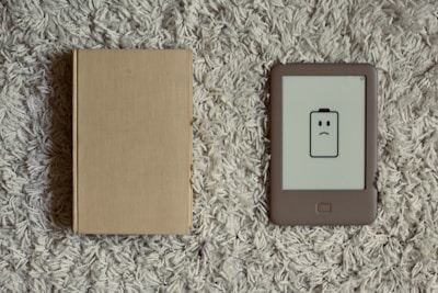 A closed beige hardcover book lies next to an e-reader with a low battery icon displaying a frowning face. Both items are placed on a textured, gray carpet.