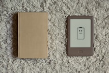 A closed beige hardcover book lies next to an e-reader with a low battery icon displaying a frowning face. Both items are placed on a textured, gray carpet.