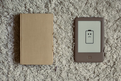 A closed beige hardcover book lies next to an e-reader with a low battery icon displaying a frowning face. Both items are placed on a textured, gray carpet.