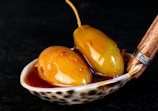 Two pickled olives with stems rest on a decorative spoon, surrounded by marinating liquid. The surface reflects light, highlighting a glossy texture. Wooden handle with metal wrapping adds an artisanal touch.