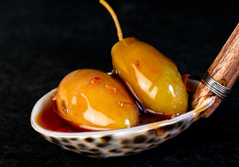 Two pickled olives with stems rest on a decorative spoon, surrounded by marinating liquid. The surface reflects light, highlighting a glossy texture. Wooden handle with metal wrapping adds an artisanal touch.