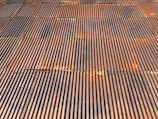 A close-up view of a corrugated metal roof with a rusted, aged appearance. The panels are aligned in a systematic pattern with alternating ridges and grooves. Various rust and weathering marks are present across the metal, adding texture and character.