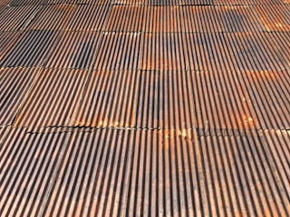 A close-up view of a corrugated metal roof with a rusted, aged appearance. The panels are aligned in a systematic pattern with alternating ridges and grooves. Various rust and weathering marks are present across the metal, adding texture and character.