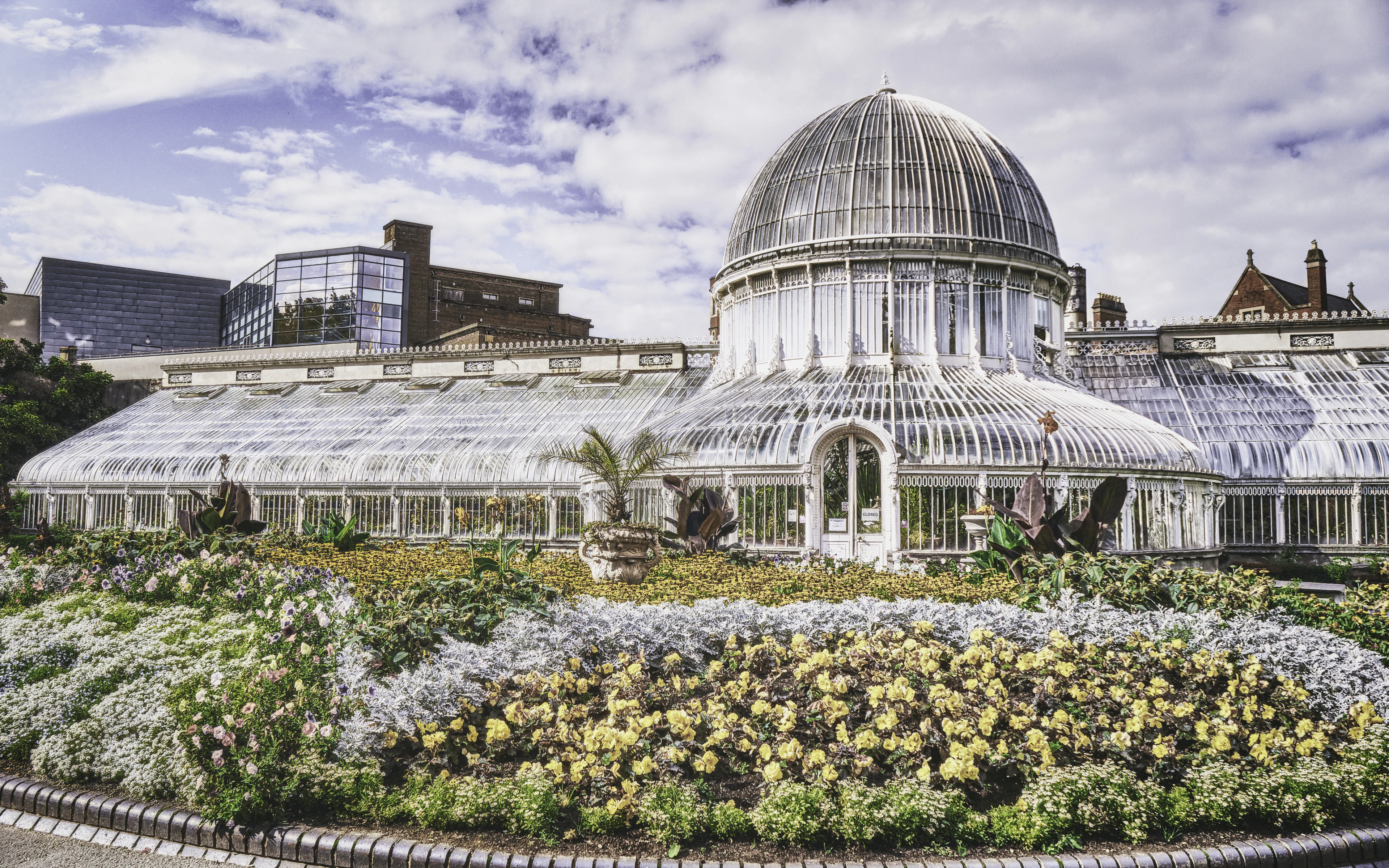 The Victorian glass Palm House at Belfast's Botanic Gardens, ringed by beds of yellow and purple flowers.