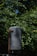 A large, weathered metal tank mounted on a rusted support pole amidst dense green foliage. The tank appears old and worn, with visible signs of chipping paint.