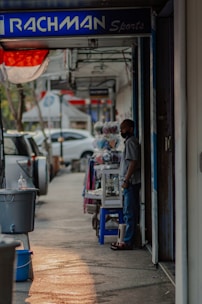 A person stands beside a shop on a street lined with parked cars. The sign above the shop reads 'RACHMAN Sports.' Nearby, various items, including clothing, are displayed on a table. The scene is shaded with a hint of sunlight casting on the sidewalk.