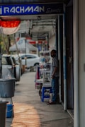 A person stands beside a shop on a street lined with parked cars. The sign above the shop reads 'RACHMAN Sports.' Nearby, various items, including clothing, are displayed on a table. The scene is shaded with a hint of sunlight casting on the sidewalk.