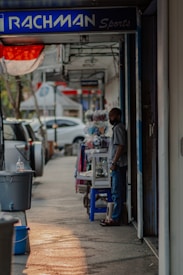 A person stands beside a shop on a street lined with parked cars. The sign above the shop reads 'RACHMAN Sports.' Nearby, various items, including clothing, are displayed on a table. The scene is shaded with a hint of sunlight casting on the sidewalk.