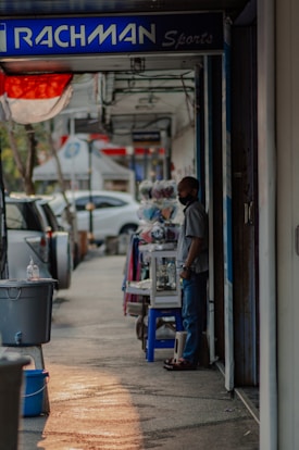 A person stands beside a shop on a street lined with parked cars. The sign above the shop reads 'RACHMAN Sports.' Nearby, various items, including clothing, are displayed on a table. The scene is shaded with a hint of sunlight casting on the sidewalk.