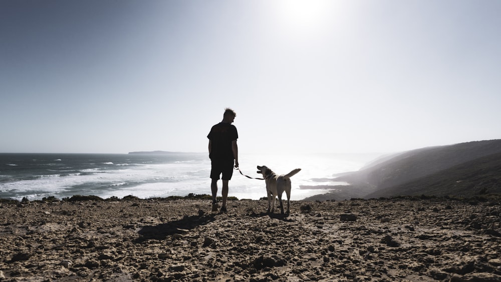 man in black jacket and brown pants standing beside brown short coated dog on seashore during