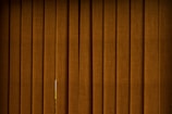 Vertical brown blinds cover a window, allowing only a slight hint of light to pass through the fabric. The slats are close together, creating a pattern with subtle texture and depth.