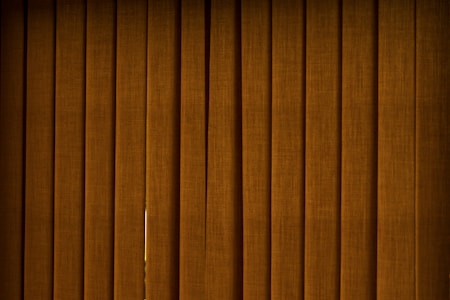 Vertical brown blinds cover a window, allowing only a slight hint of light to pass through the fabric. The slats are close together, creating a pattern with subtle texture and depth.