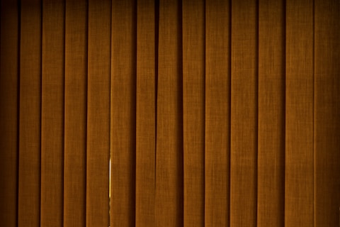 Vertical brown blinds cover a window, allowing only a slight hint of light to pass through the fabric. The slats are close together, creating a pattern with subtle texture and depth.