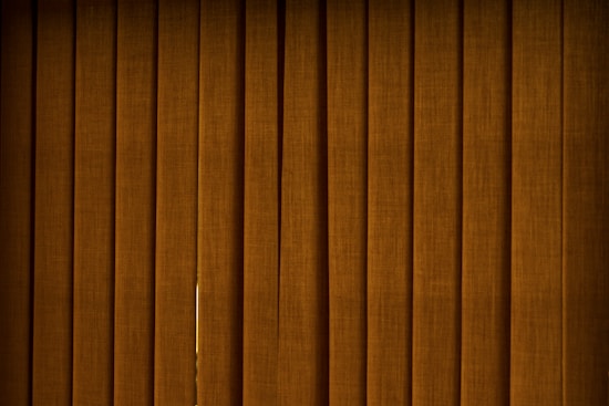 Vertical brown blinds cover a window, allowing only a slight hint of light to pass through the fabric. The slats are close together, creating a pattern with subtle texture and depth.