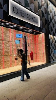 A person walks past a storefront display of a luxury store featuring mannequins. The window display is illuminated with a soft, ambient light and showcases fashion items such as bags and a red garment. The store's name is prominently displayed above the window.