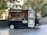 A black food truck with white accents is parked outdoors, selling handmade doughnuts. The side is open to display a variety of doughnuts on trays, with a sign indicating 'Card Only' payments. Large posters of doughnuts decorate the truck, and the leafy trees in the background suggest a park-like setting.