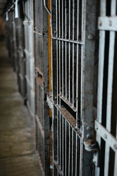 A close-up view of a row of old, rusty iron bars of prison cells. The bars are worn and weathered, with visible rust and peeling paint. There is a narrow passageway between the cells.