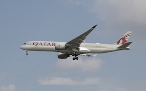 An airplane with Qatar Airways branding is in flight against a partly cloudy sky. The aircraft is a modern jetliner with a sleek design and visible landing gear.
