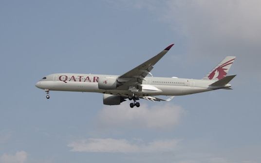 An airplane with Qatar Airways branding is in flight against a partly cloudy sky. The aircraft is a modern jetliner with a sleek design and visible landing gear.