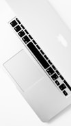 A silver laptop partially closed with a visible keyboard, trackpad, and iconic logo on the lid. The design is sleek and minimalistic, emphasizing a modern aesthetic.