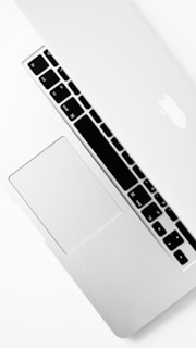 A silver laptop partially closed with a visible keyboard, trackpad, and iconic logo on the lid. The design is sleek and minimalistic, emphasizing a modern aesthetic.