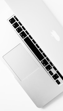A silver laptop partially closed with a visible keyboard, trackpad, and iconic logo on the lid. The design is sleek and minimalistic, emphasizing a modern aesthetic.