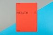 A vibrant red card with the word 'HEALTH' prominently displayed in black text, accompanied by smaller text indicating the number 09 (77) and phrases such as '77 HUMAN NEEDS' and '(physical)' at the top. The background consists of intersecting areas of blue and gray-green color.