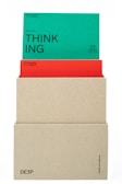 A beige box holds two cards. The top card is green with the word 'THINKING' prominently displayed in black text along with the phrase '(actional)' and other smaller text details. The card beneath is red, partially obscured by the green card, also containing textual information. The box has minimalistic text on its lower part.