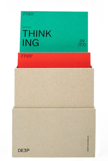 A beige box holds two cards. The top card is green with the word 'THINKING' prominently displayed in black text along with the phrase '(actional)' and other smaller text details. The card beneath is red, partially obscured by the green card, also containing textual information. The box has minimalistic text on its lower part.