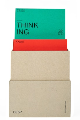 A beige box holds two cards. The top card is green with the word 'THINKING' prominently displayed in black text along with the phrase '(actional)' and other smaller text details. The card beneath is red, partially obscured by the green card, also containing textual information. The box has minimalistic text on its lower part.